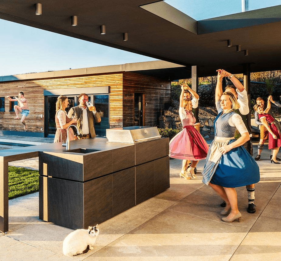 People dressed in dirndls and lederhosen dancing on a terrace. Next to them an outdoor kitchen and in the background a pool