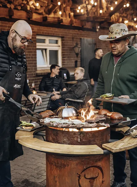 Zwei Männer grillen unterschiedlichstes Grillgut am offenen Feuer einer MOESTA Bandit Feuerstelle auf einer Terrasse