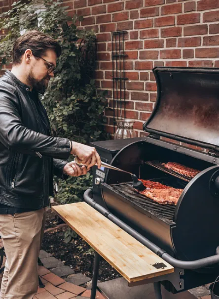 Ein Mann mit Lederjacke wendet Spare Ribs auf einem MOESTA Sheriff Pellettgrill mit Bambus Ablagebrett