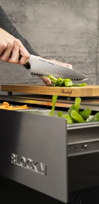 Nahaufnahme von zwei Händen, die mit einem Messer Frühlingszwiebeln auf einem Flammkraft Schneidebrett schneiden