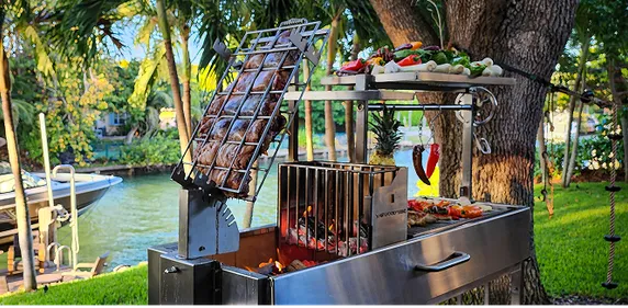 Tagwood Grill am Wasser mit geneigtem Grillrost, Gemüse und Fleisch für Grillparty vorbereitet.